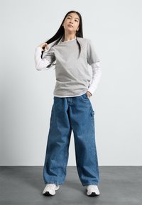Gray t-shirt layered over a white long-sleeve shirt, paired with wide-leg blue denim pants. Footwear is white sneakers with accents.