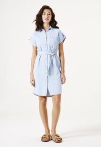 Helles blaues Jeanshemdkleid mit einer durchgehenden Knopfleiste, kurzen umgeschlagenen Ärmeln und einem Bindegürtel in der Taille. Getragen mit braunen, halb offenen Schuhen.