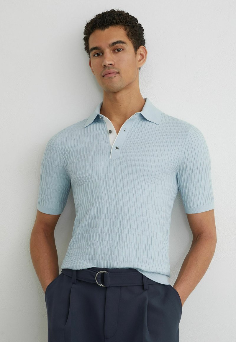 Junger Mann mit kurzen lockigen Haaren, der ein hellblaues, strukturiertes, kurzärmeliges Poloshirt und dunkle marineblaue Hosen mit Gürtel trägt.