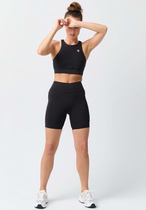 Frau in schwarzem Sport-BH und Shorts steht, hebt die Arme zur Stirn, trägt weiße Laufschuhe vor einfachem Hintergrund.