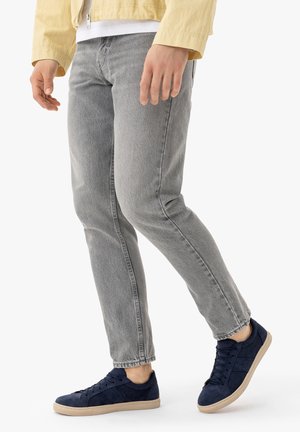 Partea inferioară a corpului unei persoane care poartă blugi gri deschis, adidași bleumarin cu talpă bej, o cămașă albă și o jachetă galbenă.