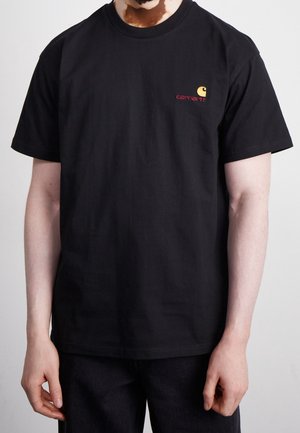 Mann trägt schwarzes Carhartt-T-Shirt mit kleinem gelb-rotem Logo auf der linken Brust, steht mit entspannten Armen an den Seiten.
