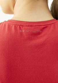 Rdeča majica s krožnim izrezom in mehkim materialom, na hrbtni strani pa je subtilno natisnjen logotip "SWEATY BETTY".
