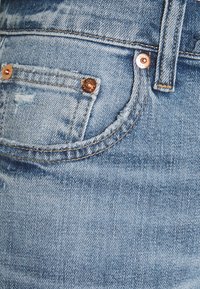 Ljusblå denimjeans nära bild som visar en frammyntficka med kopparnitar, bälteöglor och synliga sömnadsdetaljer.