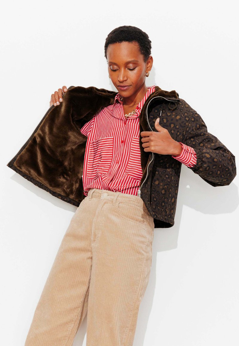 Personne portant un pantalon beige en velours côtelé et une chemise rouge à rayures, ouvrant une veste foncée à imprimé léopard avec une doublure en fourrure marron.