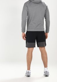 Haut à capuche gris avec une finition texturée, associé à un short noir présentant un panneau gris foncé. Des chaussures de sport complètent la tenue.