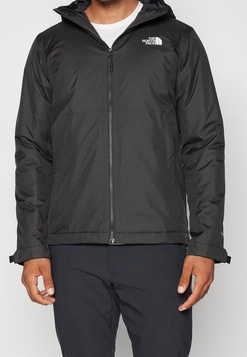 Homme portant une veste à capuche noire North Face zippée, associée à un pantalon noir, debout les bras détendus le long du corps.