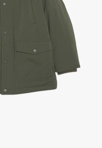 Veste isolante vert olive dotée d'une fermeture éclair frontale, de boutons-pression et d'une poche poitrine. Les poignets côtelés ajoutent de la texture au design.