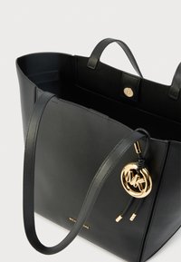 Sac cabas en cuir noir avec deux poignées, ferrures dorées et pendentif logo. Intérieur spacieux avec petite poche et fermeture à bouton.