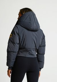 Giacca puffer grigia scura accorciata con un grande cappuccio, dotata di chiusura con zip, orlo a costine e una patch con logo sulla manica.