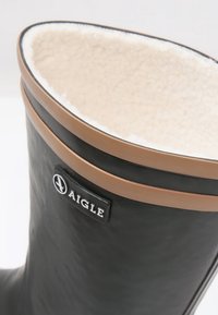 Botte en caoutchouc noire avec une bordure marron et une doublure douce de couleur crème. Présente une étiquette logo sur le côté et un extérieur lisse et texturé.