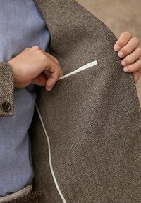 Brown herringbone patterned jacket with beige inner lining; white accent piping along the pocket; hand reaching into the inner pocket.