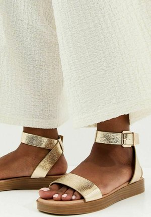 Sandalias planas doradas brillantes con tiras en el tobillo y hebillas, usadas con pantalones blancos de pierna ancha y textura, en piel oscura.