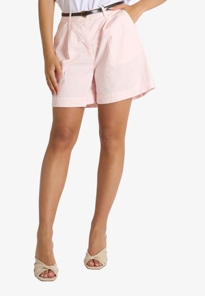 Donna che indossa pantaloncini rosa chiaro a vita alta con cintura nera, top bianco e sandali beige con tacco e punta aperta, posando con una mano in tasca.