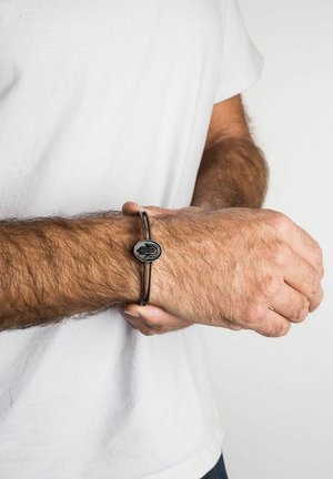 Pulsera de cuero negro con un medallón ovalado de plata, que presenta una superficie texturizada y un diseño grabado, llevada en una muñeca desnuda.