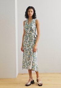 Robe portefeuille fleurie avec une base blanche ornée de motifs floraux verts et jaunes, mi-longue, en tissu texturé, associée à des sandales à plateforme noires.