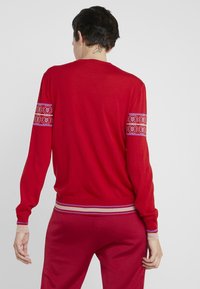 Suéter rojo de punto con una banda decorativa en tonos rosa y blanco en las mangas y un dobladillo a rayas. Textura suave y diseño ajustado.