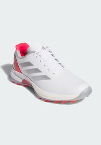 Witte sportieve schoen met rode accenten. Glad synthetisch bovenwerk, gevoerde grijze kraag en een gestructureerde zool met roze spikes voor grip.