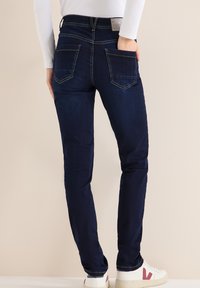 Jean skinny bleu foncé avec une texture en denim lisse. Comprend une taille haute, cinq poches, et des coutures jaunes. Baskets blanches avec des accents.