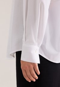 Camicia bianca a maniche lunghe realizzata in tessuto liscio; presenta un polsino con bottone e un orlo leggermente curvo, su uno sfondo nero texturizzato.