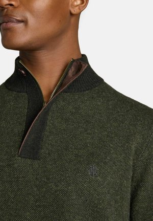 Nahaufnahme einer Person, die einen dunkelgrünen strukturierten Pullover trägt, mit einem teilweise geöffneten schwarzen Rippenkragen und einem kleinen gestickten Logo auf der Brust.