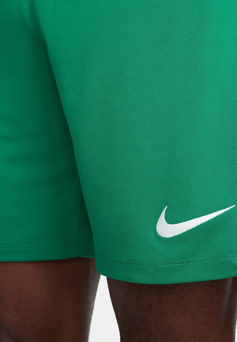 Shorts de sport verts avec logo Nike Swoosh blanc sur la jambe inférieure droite, portés par une personne à la peau foncée.