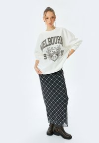 Sudadera blanca de gran tamaño con estampado de "Melbourne", combinada con una falda larga de cuadros en tonos oscuros y botas de tacón grueso marrones.
