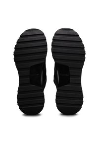 Semelles de chaussures noires avec des rainures horizontales profondes et la marque "Calvin Klein" visible près de la zone de la voûte plantaire sur fond blanc.