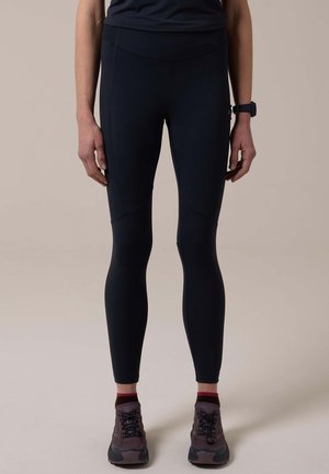 Persoon draagt zwarte strakke legging, donkerrode enkelsokken, paarse en zwarte hardloopschoenen en een zwart horloge, staand tegen een effen achtergrond.