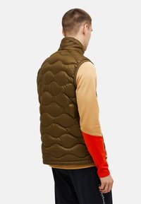 Olivgrüwestuine Weste mit wellenförmiger Steppstruktur, hohem Kragen und ärmellosem Design. Getragen über einem hellbraunen und orangefarbenen Langarmshirt.