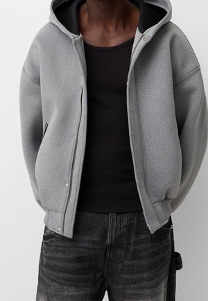 Persoon draagt een grijze zip-up hoodie met een zwarte binnenvoering, een zwarte mouwloze top en vervaagde zwarte spijkerbroek tegen een effen achtergrond.