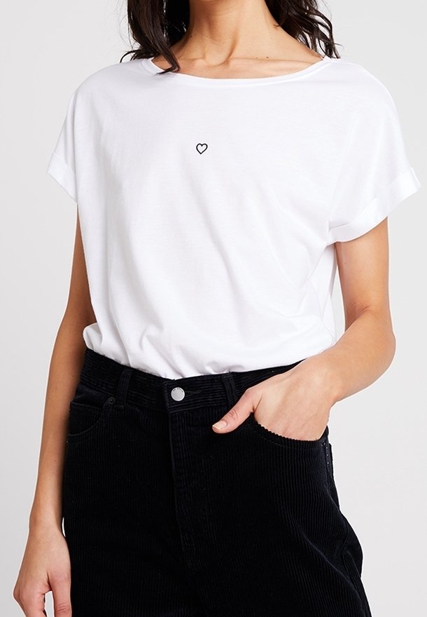 Hvid kortærmet t-shirt med rund halsudskæring, der har et lille sort hjerte broderet på brystet, parret med sorte corduroybukser.