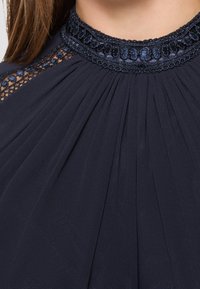 Tmavě modré šaty s sklady, zahrnující texturovaný krajkový detail na krku a složitou výšivku podél límce pro dodatečný design.