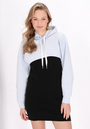 Junge Frau mit langen blonden Haaren, die ein schwarzes und hellgraues Kapuzen-Sweatshirtkleid trägt, stehend vor einem einfarbigen Hintergrund.