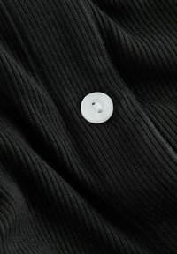 Tissu noir côtelé avec un bouton en plastique blanc, présentant un trou central et une légère texture, étalé à plat avec des plis doux.