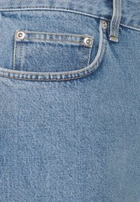 Světle modrá džínovina má zadní kapsu se stříbrným kováním a kontrastním prošitím. Textura je hladká s mírně opotřebovaným vzhledem.