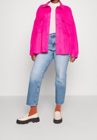 Camisa chaqueta de gran tamaño en fucsia con dos bolsillos delanteros, suéter blanco de canalé, jeans rectos de mezclilla azul claro y mocasines beige con suelas negras.
