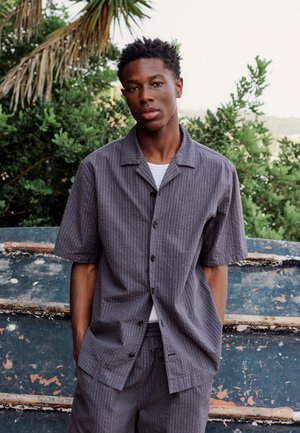 Jeune homme debout à l'extérieur près des arbres et d'un bateau usé, portant une chemise grise à manches courtes boutonnée et un short assorti.