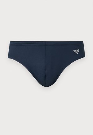 Pánske plavky tmavomodrej farby s nízkym pásom a bielym vyšitým logom na pravej prednej strane na jednofarebnom pozadí.