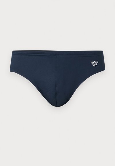 Ανδρικό μαγιό slip σε ναυτικό μπλε με χαμηλή μέση και λευκό κεντημένο λογότυπο στη δεξιά μπροστινή πλευρά, σε μονόχρωμο φόντο.