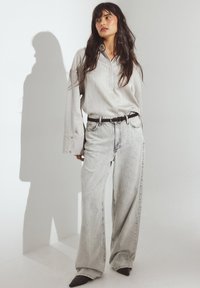 Camisa de manga larga gris claro con tapeta y puños anchos, combinada con jeans gris claro de corte suelto. El modelo lleva un cinturón negro y zapatos de punta.