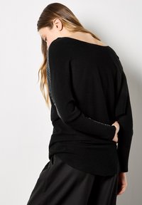 Donna con capelli lunghi che indossa un maglione nero con dettagli bianchi sulle maniche, in piedi di lato con una mano dietro la schiena.