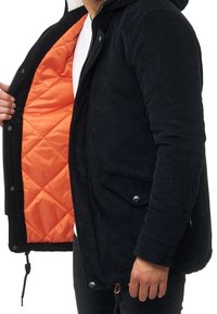 Person trägt eine schwarze Cordjacke mit orangefarbigem Steppfutter und hält die Jacke offen, um das Innere zu zeigen, vor einem weißen Hintergrund.