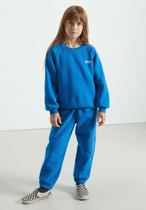 Vaikas su ilgais plaukais, apsirengęs mėlynu džemperiu ir derančiomis kelnėmis, stovi ant šviesiai pilko grindų, apsivilkęs šachmatų rašto šlepetes.