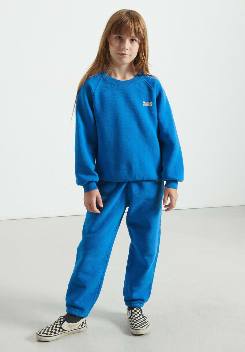 Bērns ar garām matiem, tērpies zilā džemperī un atbilstošās biksēs, stāv uz gaiši pelēka grīdas, uzvilcis pārbaudītas slip-on kurpes.