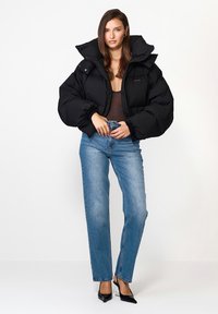 Czarna kurtka puchowa z wysokim kołnierzem i pikowanym wzorem, zestawiona z niebieskimi dżinsami o wysokim stanie i czarnymi butami na szpilkach.