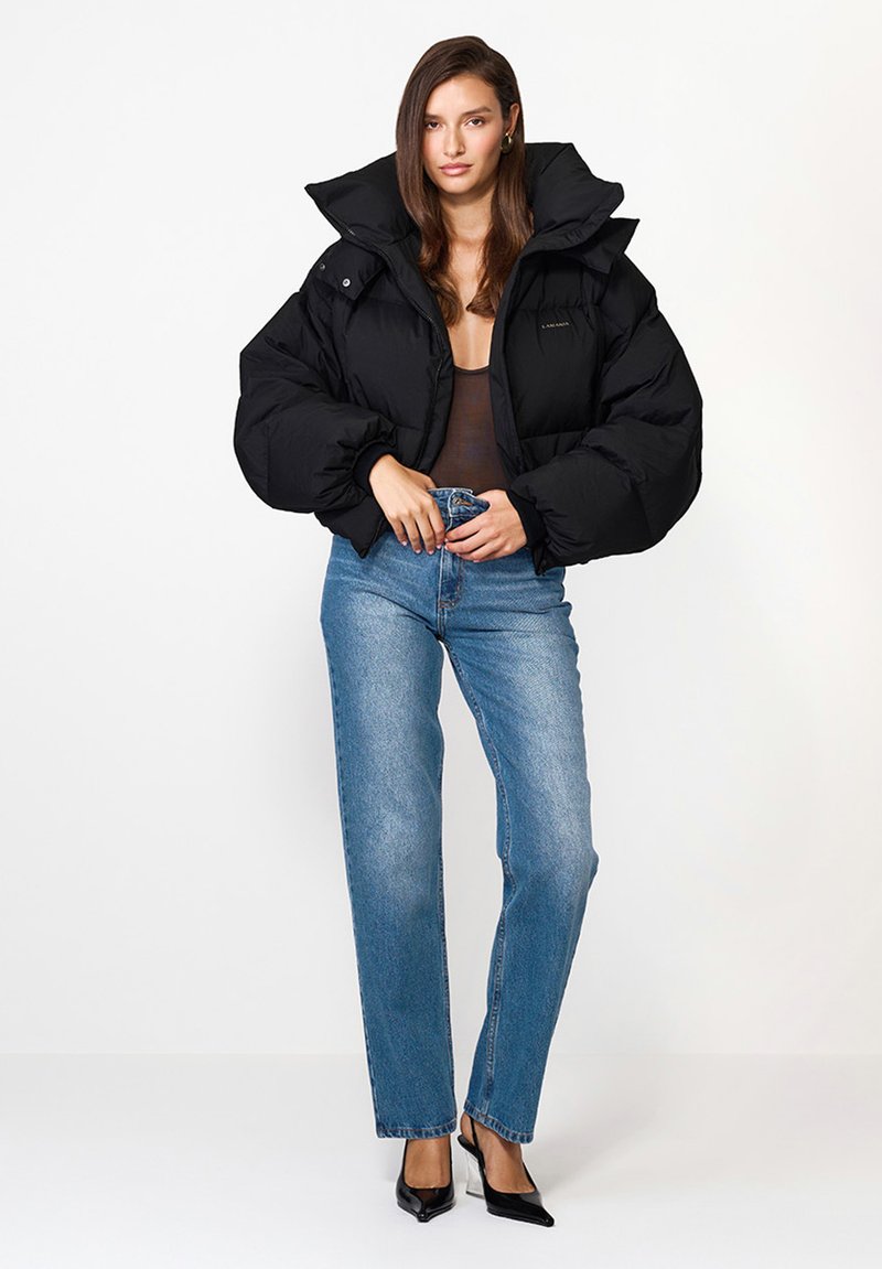 Czarna kurtka puchowa z wysokim kołnierzem i pikowanym wzorem, zestawiona z niebieskimi dżinsami o wysokim stanie i czarnymi butami na szpilkach.
