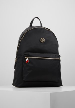 Sac à dos noir en nylon avec poche avant zippée, fermeture éclair dorée, poignée supérieure et emblème rond avec logo au-dessus de la poche sur fond uni.