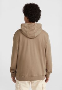 Personne aux cheveux bouclés foncés portant un sweat à capuche marron et un pantalon cargo beige clair, vue de dos sur un fond blanc uni.