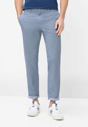 Uomo con pantaloni slim fit azzurri chiari con risvolti, scarpe da ginnastica bianche con accenti blu e camicia blu navy, in piedi su sfondo bianco.